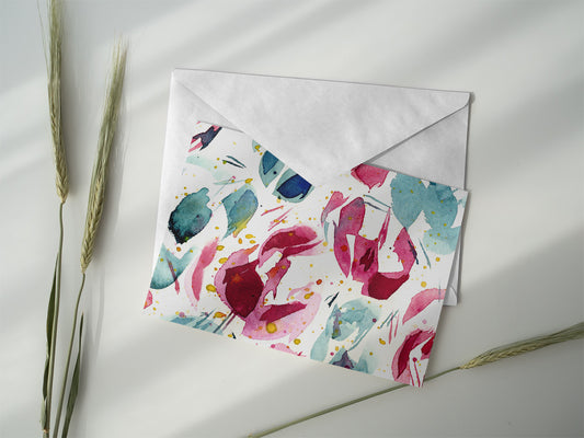 Product photo of abstract watercolor greeting card titled "Joy and Sorrow" with various blue, green, and red hues on a clean, white card. The card is laying on top of an envelope on a white background next to greenery. 